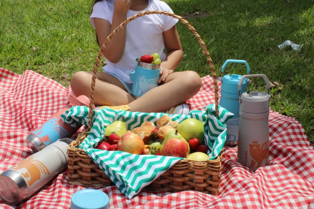 Uma criança sentada atrás de uma cesta repleta de frutas, com produtos Stanley da linha Kids ao redor dela representando a lancheira infantil térmica com itens Stanley