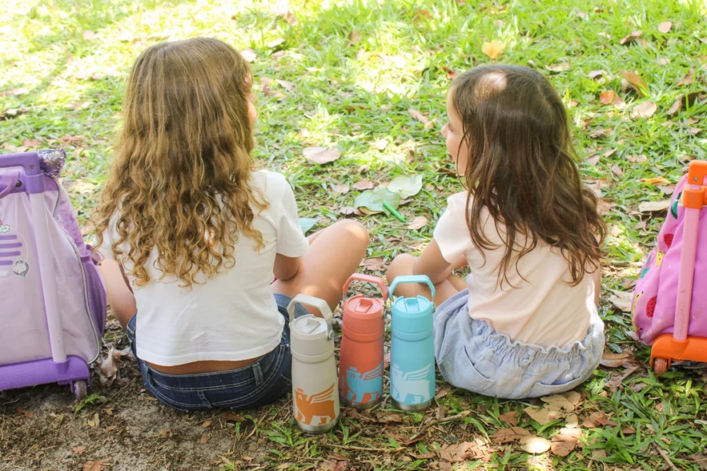Duas crianças sentadas com garrafas infantis por perto para manter a hidratação