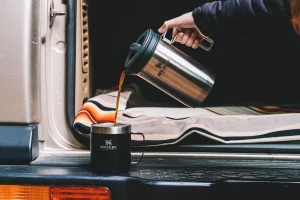 Uma pessoa usando o carro como suporte para encher uma caneca da Stanley com café usando a Prensa Francesa da Stanley para isso, mostrando que ela está em movimento por ter diversos destinos para conhecer