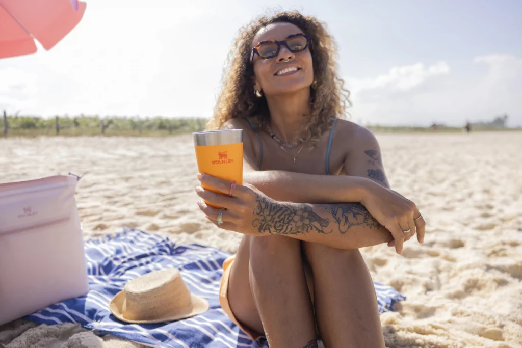 Uma mulher sentada na praia segurando o novo copo da coleção Stanley Heat Wave